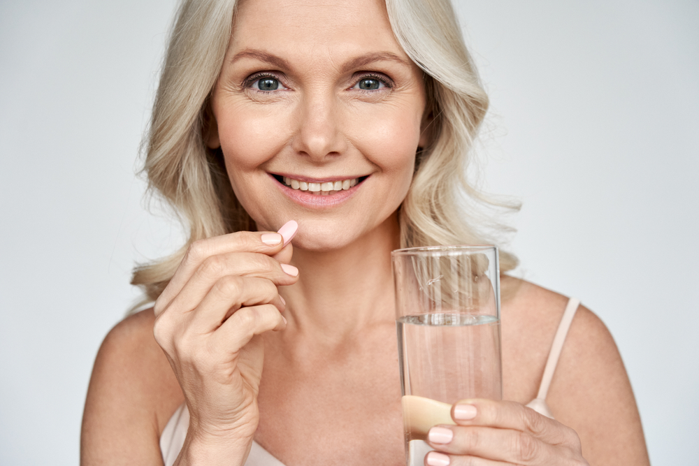 older smiling woman holding a glass of water and a supplement