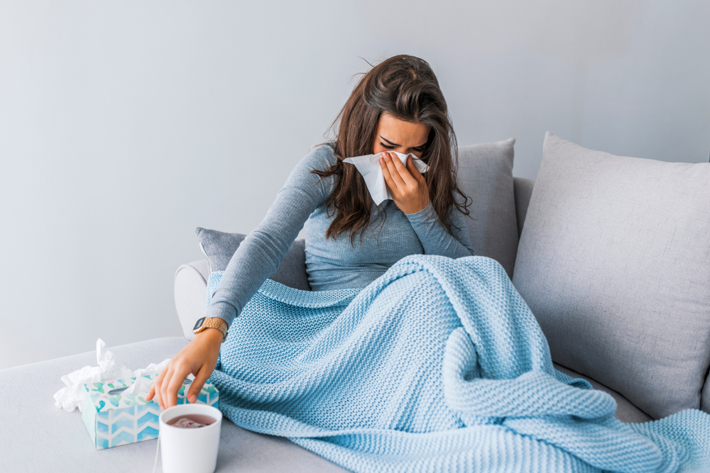 woman blowing her nose into a tissue while reaching for tea covered in a blanket on the couch